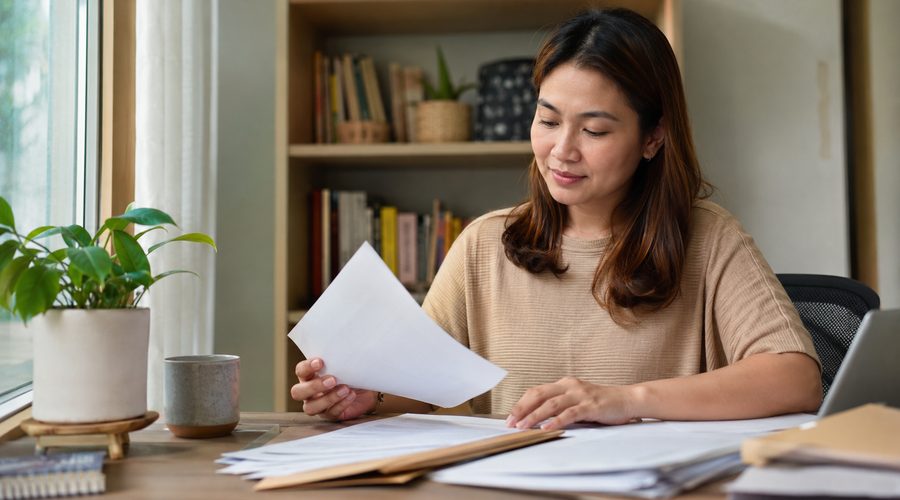 A woman reviewing paperwork at a home desk beside a stack of files, getting SSS, PhilHealth, and Pag-IBIG contributions in order